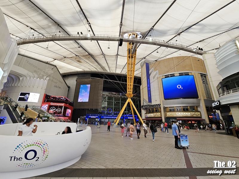 The O2 內部中庭廣場，中央為黃色支架結構與圓頂攀登設施，人群在商店與餐廳間穿梭。
