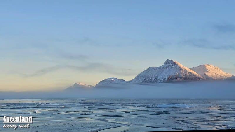 格陵蘭冰峽灣風景，前景是海面上的浮冰，遠方雪山在晨光映照下呈現粉橙色